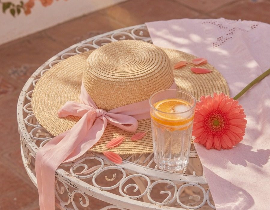 Mesa de un patio español al sol: vaso de agua con rodajas de naranja, sombrero de paja y una flor rosa